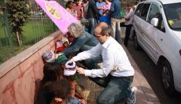 Sunny und der Fahrer weisen nun die Kinder an, sich in einer langen Reihe am Boden auf dem Gehsteig hinzusetzen. An dem dahinter befindlichen Zaun befestigen wir den Friendcircle-Worldhelp- Banner und Peter und Christian verteilen direkt Mützen.