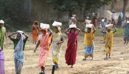 Nachmittags werden Steine und Ziegel für die Mauer geliefert. Der Baubeginn wird traditionell mit einer Zeremonie gefeiert, bei der die Frauen der Kolonie ihre besten Gewänder anziehen und die schweren Steine zum Graben für das Fundament bringen.